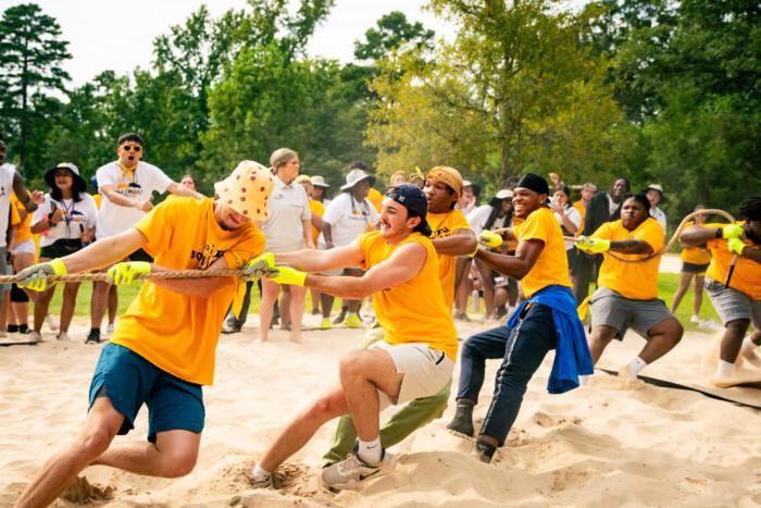 Students play tug-of-war on sand, wearing gold shirts. The audience cheers in the background.